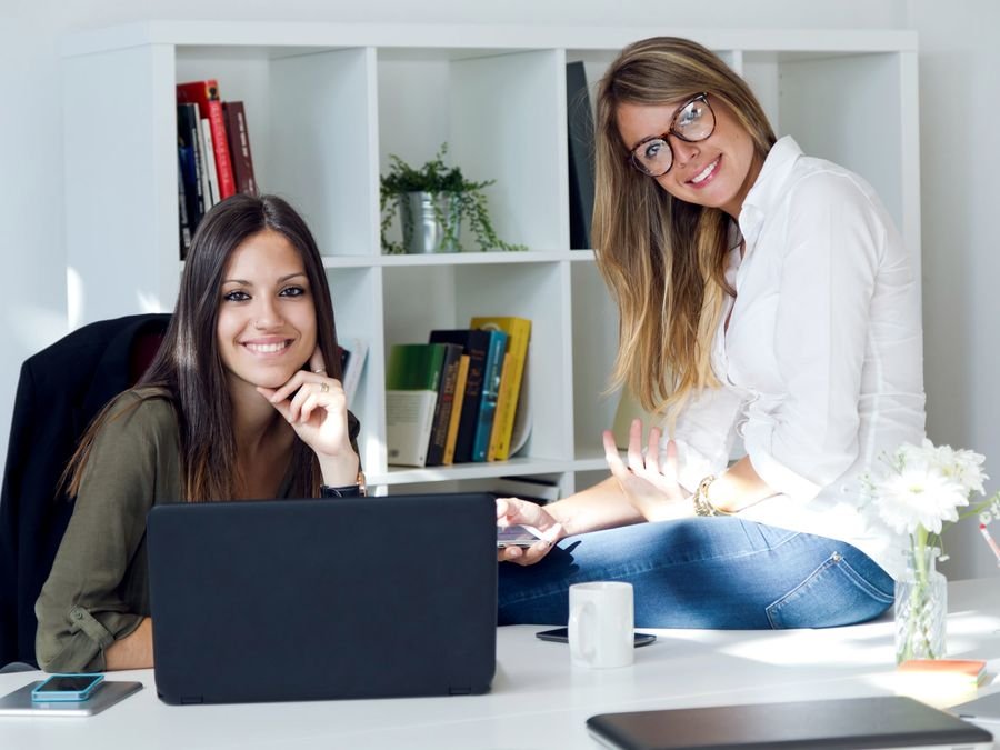 Duas sócias sorrindo no escritório, ao lado de um notebook